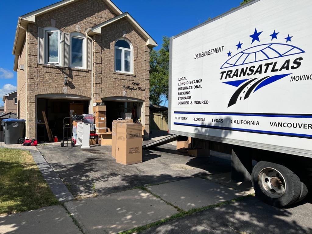 Moving truck labeled Transatlas parked in driveway while boxes and furniture are loaded from a two-story brick house with open garages nearby.