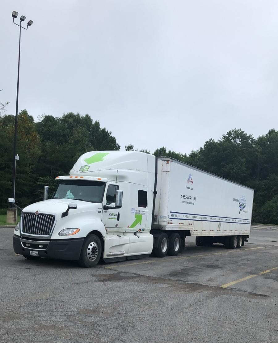 Front-right view of a white semi-truck with a trailer parked in a large empty lot, trees in the background and a tall street light nearby.