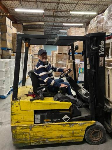 Man wearing a striped sweater and cap operating a yellow Yale forklift in a warehouse, with tall stacks of boxes and pallets around him.