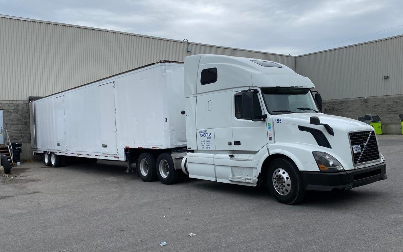 White semi tractor-trailer with a long white trailer parked in a loading yard beside an industrial building plus a few green dumpsters in the background.
