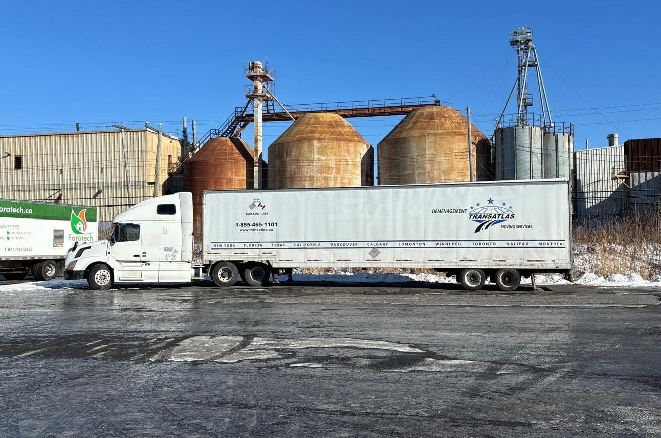 White semi-truck with a long trailer parked beside rusted industrial silos under a clear blue sky in an industrial yard, with snow on the ground.