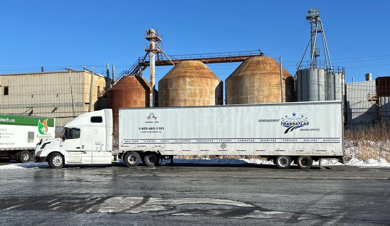 White semi-truck with a long cargo trailer parked beside rusted industrial silos under a clear blue sky.