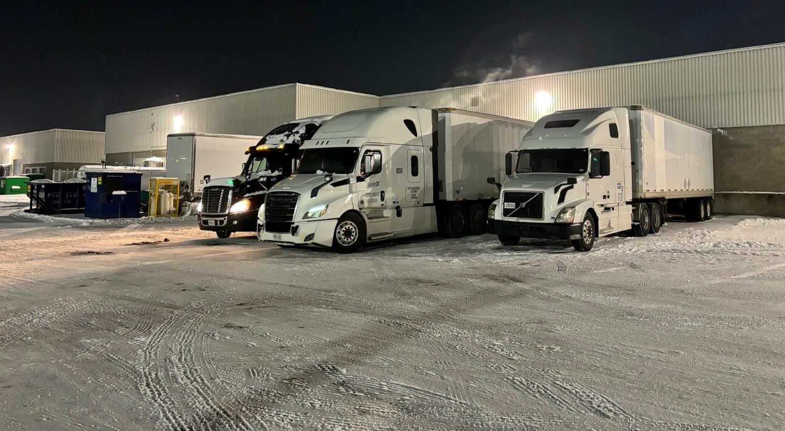 Night scene of three white semi-trucks with trailers parked in a snow-covered lot beside a large warehouse building, illuminated by yard lights.